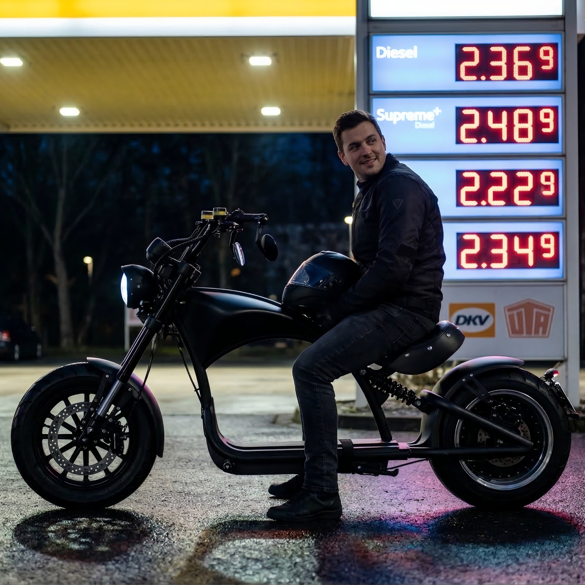Ein Mann sitzt auf einem mattschwarzen Star-Biker E-Chopper M1P Custom vor einer Preisanzeige an einer Tankstelle bei Nacht – Symbol für Elektromobilität als günstige Alternative zu hohen Benzinpreisen.
